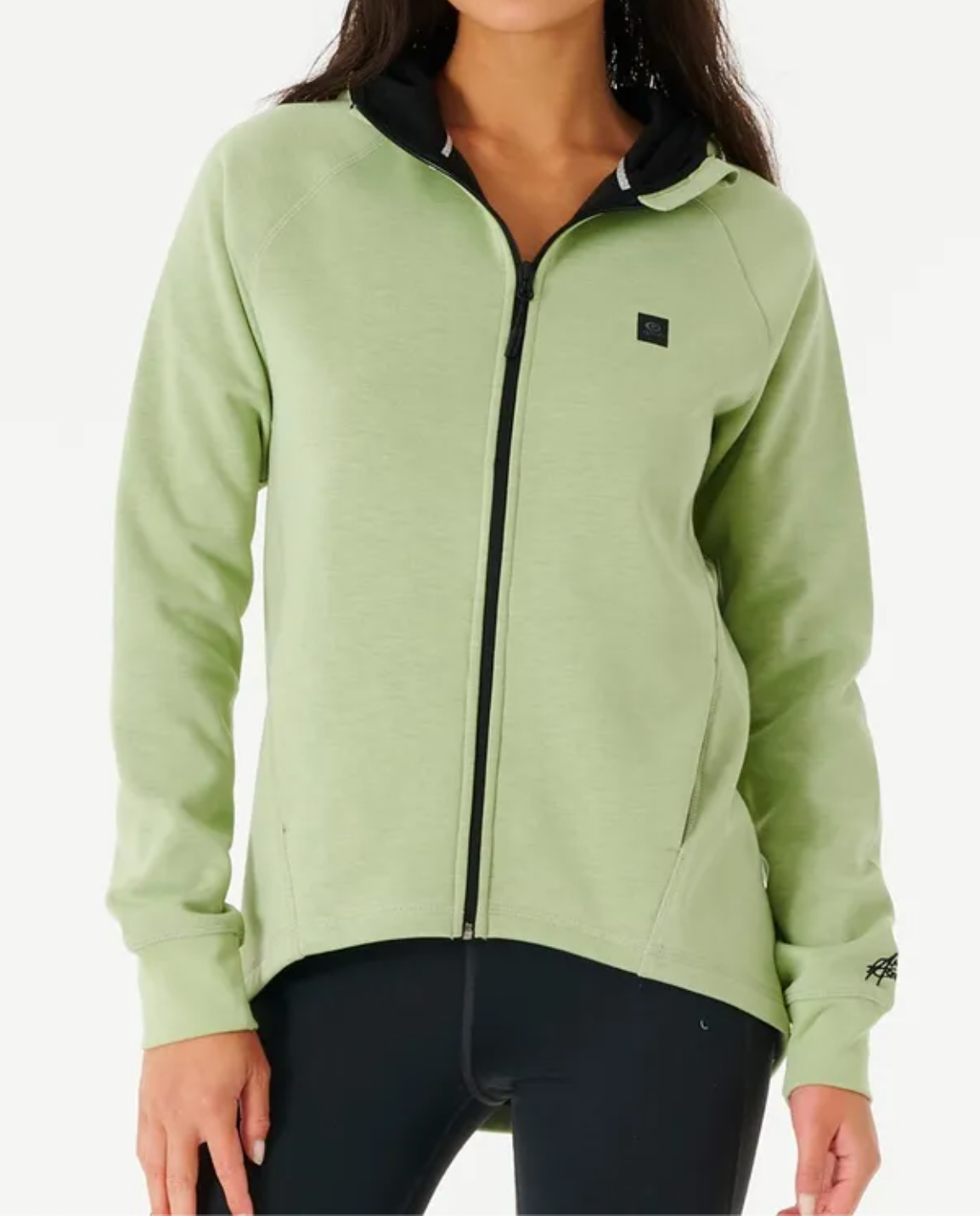 A woman wears a light green Rip Curl Anti Series Flux II Zip hoodie with black hood lining and black leggings, featuring a small black logo on the chest. Ideal for winter, she stands confidently against a plain white background.
