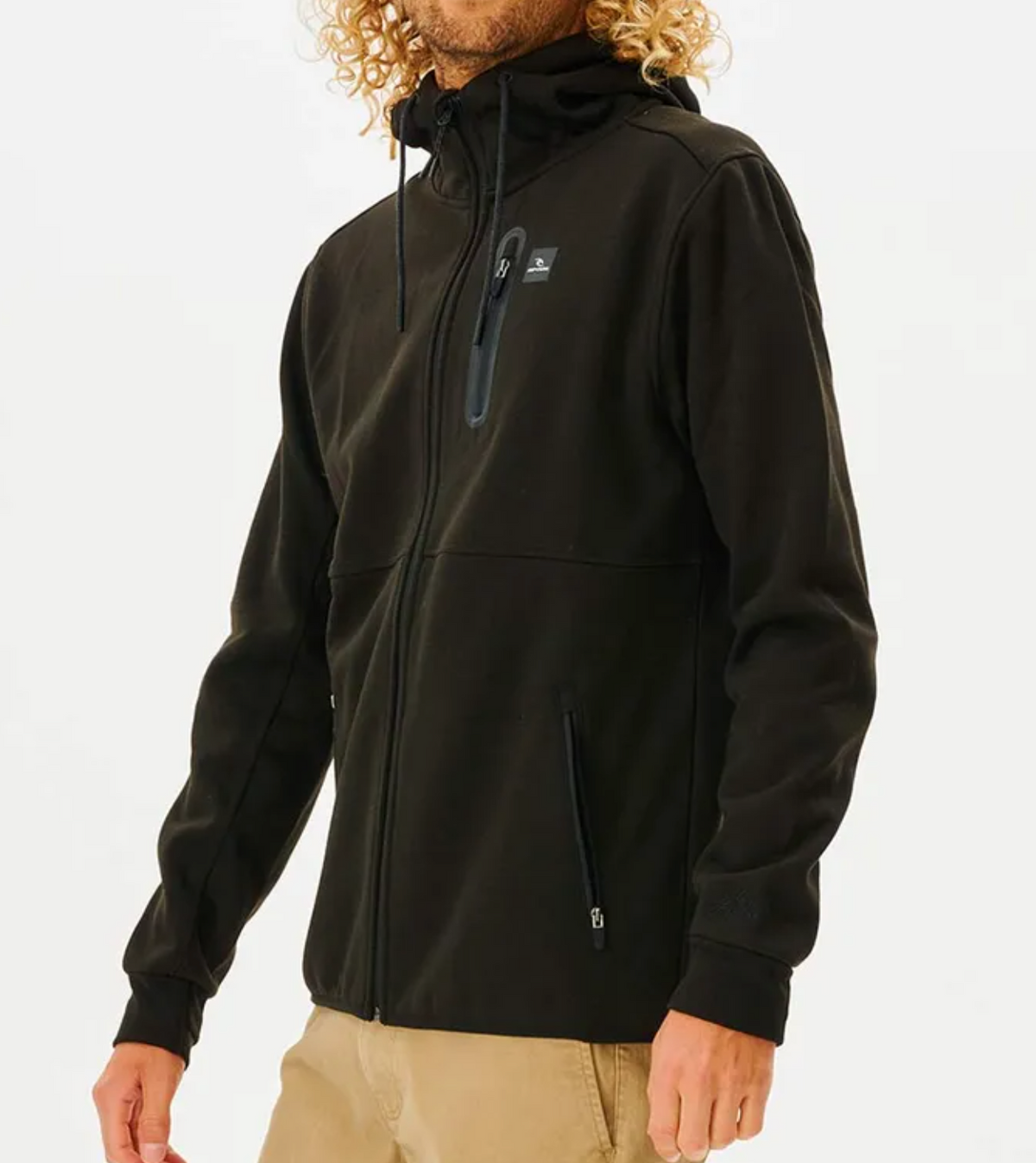 Wearing a black Rip Curl Anti Series Departed Zip Thru hoodie with a chest pocket, someone stands against a white background. With curly hair and beige pants, this durable water-resistant jacket features zippered side pockets, ideal for winter clothing.