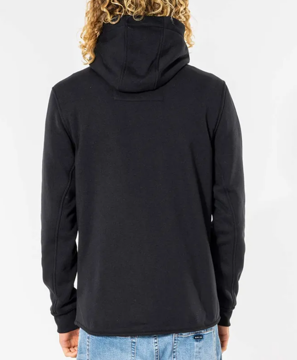 A person with long curly hair, draped in a black Rip Curl Anti Series Departed Zip Thru and blue jeans, stands facing away against a plain white background.