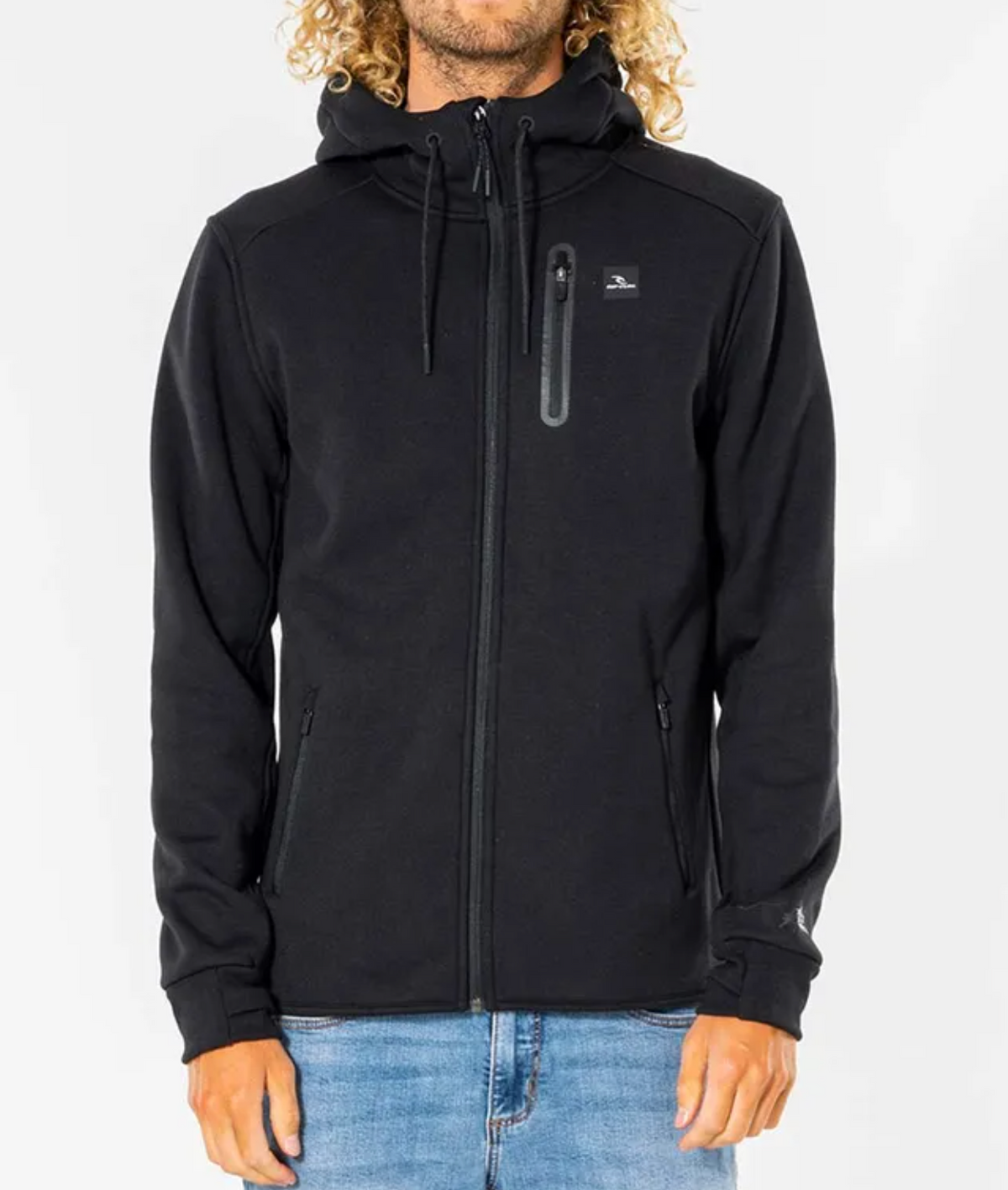 A person wearing the Rip Curl Anti Series Departed Zip Thru, a durable black hoodie with a hood, small chest logo, and two zippered pockets, stands against a white background in light blue jeans. This water-resistant hoodie is ideal for winter wear.