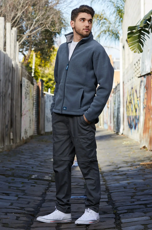 A man stands in a narrow alley adorned with graffiti, sporting the Rainbird Lumber Jacket, dark cargo pants, and white sneakers. With one hand tucked into his pocket, he gazes off into the distance. The alley is flanked by wooden fences and concrete walls.