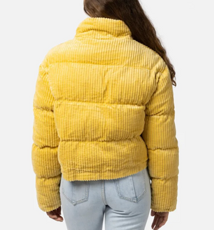 Rear view of a person with long hair wearing the Rip Curl Trippin Cord Jacket in yellow and light blue jeans. This stylish and cozy corduroy puffer jacket has a high collar and horizontal stitching, set against a plain white background.