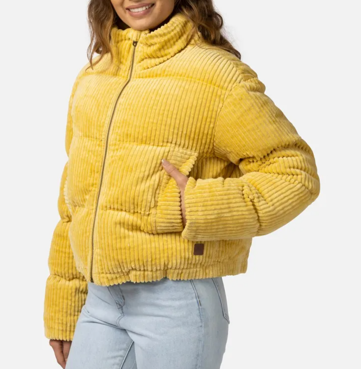 A person dons the stylish Rip Curl Trippin Cord Jacket, featuring large pockets and a yellow corduroy design, paired with light blue jeans. Smiling with one hand in a pocket, they stand against a plain background, effortlessly showcasing their comfortable jacket choice.