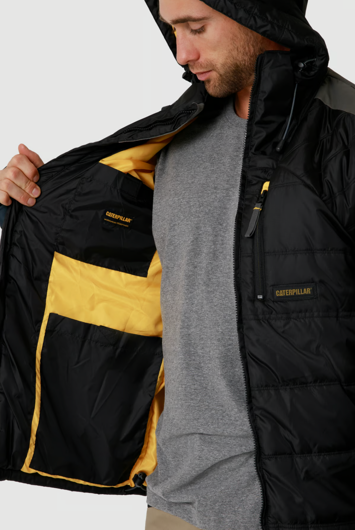 A person wears a CAT Triton Insulated Puffer Jacket, showcasing its storm blocker technology and visible yellow/black interior with inside pockets. Paired with a grey shirt underneath, the windproof fabric perfectly complements the "Caterpillar" branding.