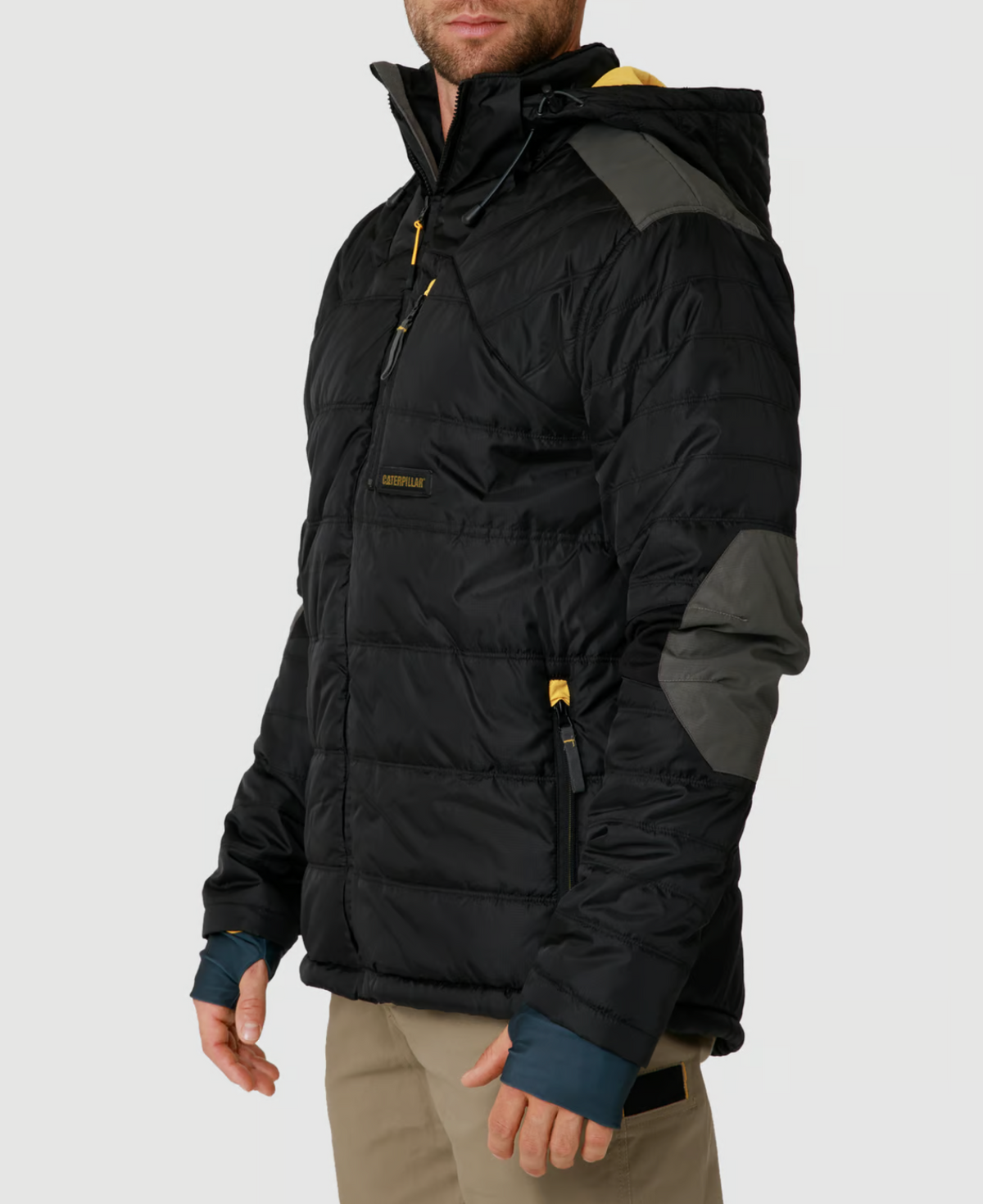 A person stands against a plain light gray background wearing the CAT Triton Insulated Puffer Jacket, which is black with gray elbow patches and yellow zippers. This windproof piece with storm blocker technology pairs perfectly with their beige pants and features a functional hood.