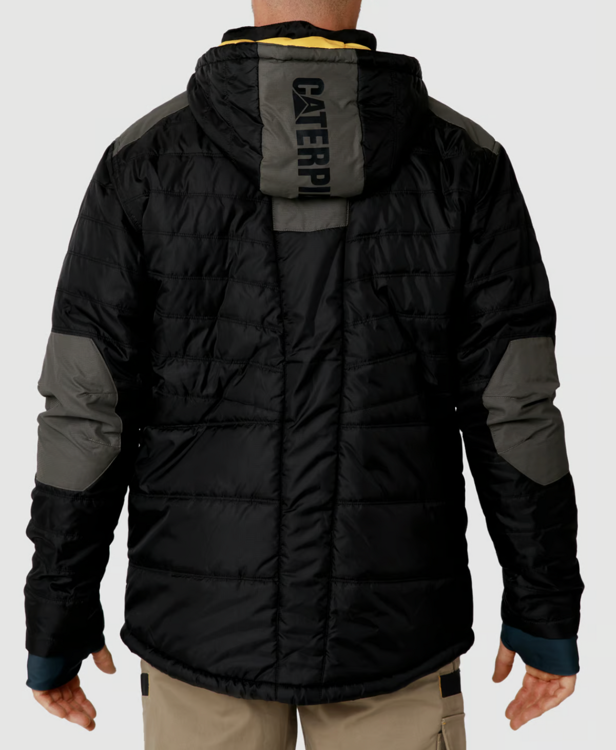 A person wearing a CAT Triton Insulated Puffer Jacket, featuring storm blocker technology and a hood, stands with their back to the viewer. The black and gray jacket has "CATERPILLAR" printed on the back, paired with khaki pants.