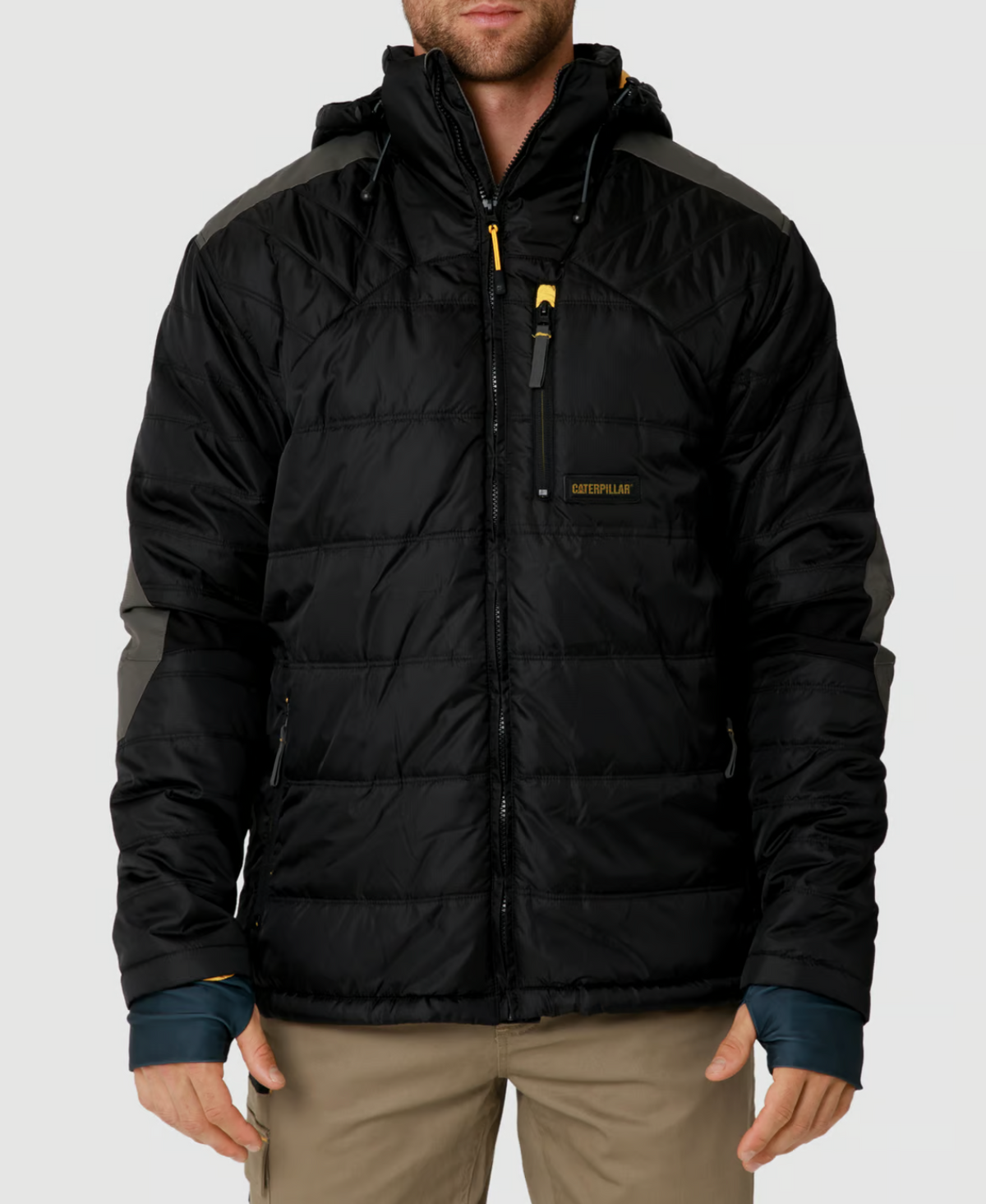 A person is wearing the CAT Triton Insulated Puffer Jacket in black with gray accents and a hood. Made from windproof fabric, it has a zippered chest pocket with a yellow pull tab and storm blocker tech. It features a logo on the chest. The person pairs it with beige pants.