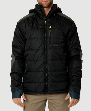 A person is wearing the CAT Triton Insulated Puffer Jacket in black with gray accents and a hood. Made from windproof fabric, it has a zippered chest pocket with a yellow pull tab and storm blocker tech. It features a logo on the chest. The person pairs it with beige pants.