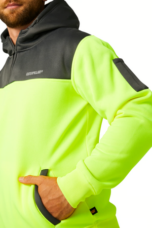 A man confidently stands in a neon yellow and gray CAT Hi Vis Hoodie, proudly displaying the "Caterpillar" branding. The hoodie features a zippered pocket on the left sleeve and his left hand comfortably rests in the front pocket. Its rugged dependability is evident with its fleece lining for added comfort, perfectly complemented by his brown beard and visible hood.