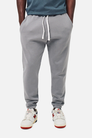 A person wearing Industrie The Del Sur Track Pant in gray with an elastic drawstring waist, a teal shirt, and white sneakers accented in red and blue stands with hands in pockets against a plain background.