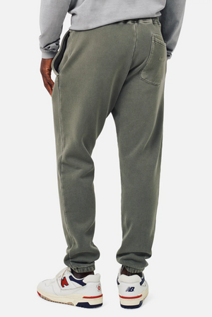 A person in a gray sweatshirt and Industrie The Del Sur Track Pant, featuring olive green color and an elastic drawstring waist, stands facing away. With hands in pockets and donning white athletic shoes accented with red and blue, they stand against a plain white background.