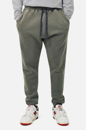 A person in Industrie The Del Sur olive green joggers with an elastic drawstring waist, paired with white sneakers accented in red and navy, stands hands-in-pockets against a white backdrop.