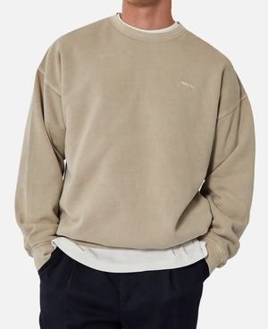 A person is shown from shoulders to waist wearing the Industrie The Del Sur Sweat, an oversized fit beige jumper over a white shirt with subtle chest text. It is paired with black pants against a plain white background.