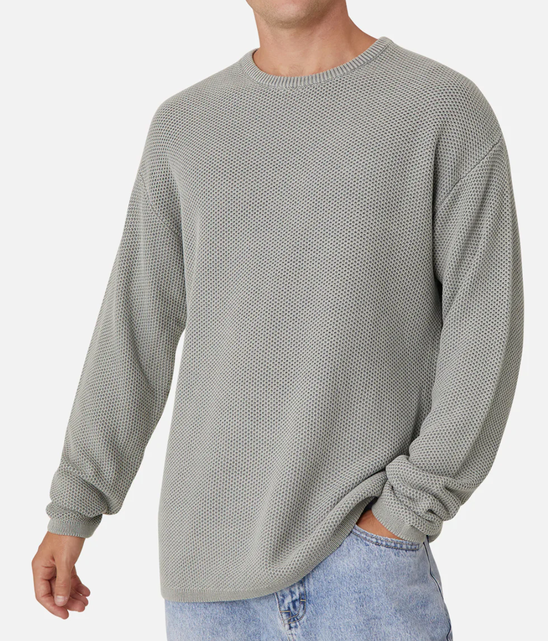A person models the Industrie The Washed Culver Knit, a 100% cotton light gray sweater with long sleeves and a classic crew neck, paired with blue jeans. They stand against a plain white background, one hand in their pocket, without showing their face.