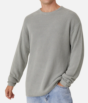 A person models the Industrie The Washed Culver Knit, a 100% cotton light gray sweater with long sleeves and a classic crew neck, paired with blue jeans. They stand against a plain white background, one hand in their pocket, without showing their face.
