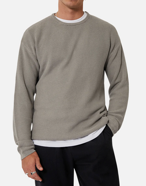 A person is clad in the Industrie The Washed Culver Knit, a comfy gray sweater made of 100% cotton, layered over a white shirt. They pair it with black pants and stand against a plain background, casually tucking their left hand into their pocket.