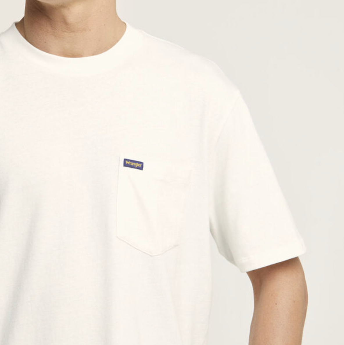 A person is wearing a Wrangler Classic Pocket Tee, featuring a plain white design with a small chest pocket and a tiny rectangular label. The image is cropped at the neck and mid-arm, highlighting the simplicity of this 100% cotton shirt.