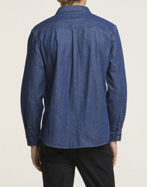 Rear view of a person in a Wrangler Roadside Denim Shirt, regular fit with long sleeves, paired with black pants. The blue denim shirt is 100% cotton, featuring a collar and buttoned cuffs against a plain white background.