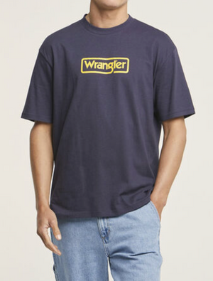 A person wears a dark blue Wrangler Border Tee with a bold yellow logo across the chest, paired with light blue jeans. They casually have one hand in their pocket as the plain white background highlights their regular fit ensemble.