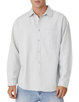 A man is wearing Industrie The Portland Shirt, a light gray relaxed-fit overshirt with long sleeves and a chest pocket, paired with light blue jeans. The casually styled cotton-linen shirt complements his laid-back look against a plain white background.