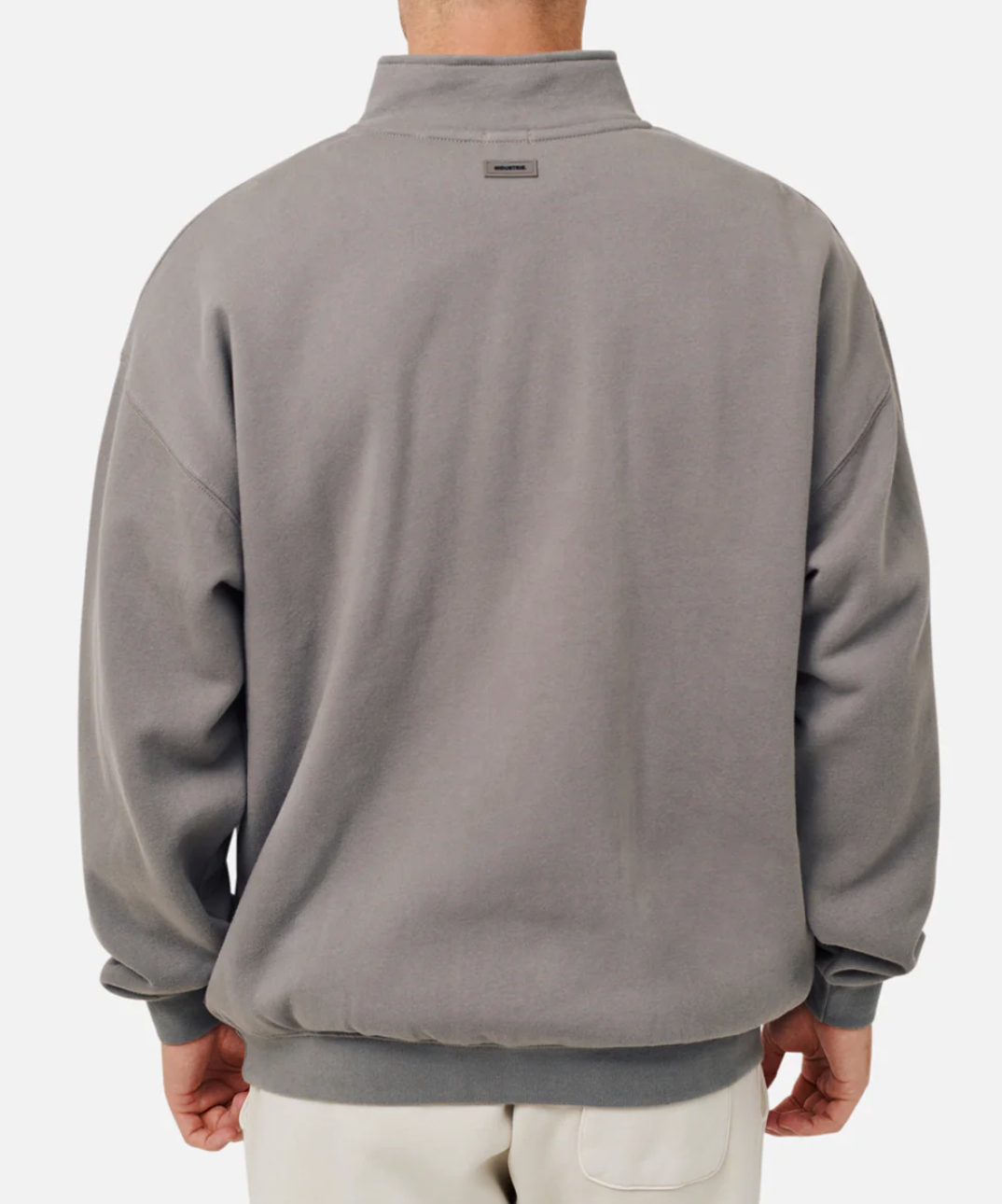 The individual wears an oversized Industrie The Del Sur Track Top, characterized by a high collar, slightly dropped shoulders, and ribbed cuffs, all in gray. Paired with light-colored pants, the ensemble projects a casual sweat vibe against a plain white background.
