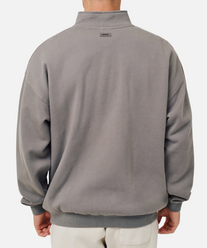 The individual wears an oversized Industrie The Del Sur Track Top, characterized by a high collar, slightly dropped shoulders, and ribbed cuffs, all in gray. Paired with light-colored pants, the ensemble projects a casual sweat vibe against a plain white background.