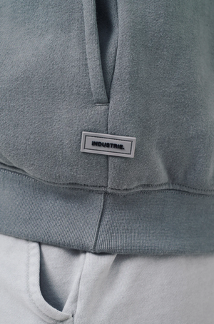 Close-up of a person wearing the Industrie The Del Sur Track Top, an oversized muted green hoodie with soft fleece lining. It features a small rectangular "INDUSTRIE" label and ribbed cuffs, paired with light gray pants.