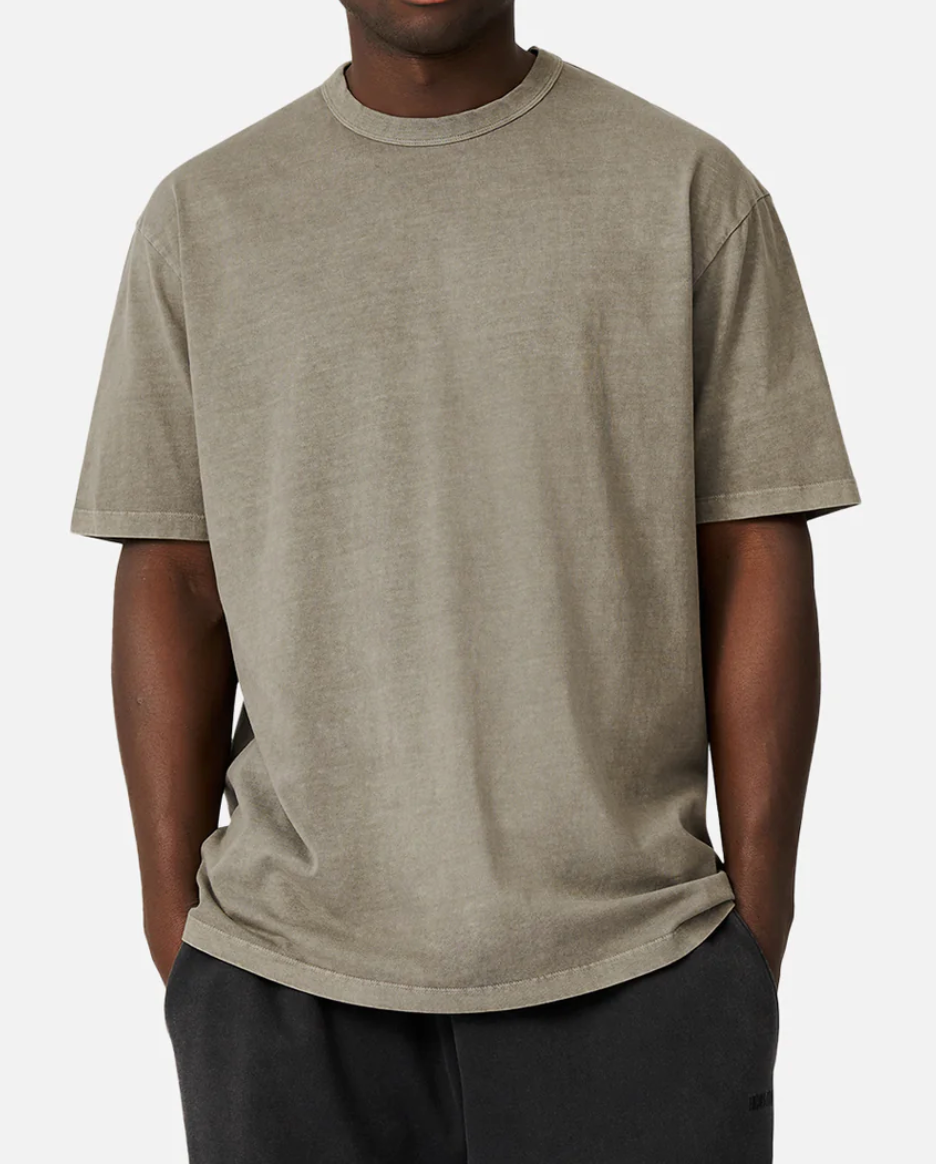 Someone wearing an oversized faded olive green Industrie The Del Sur Tee stands against a plain background, with hands partially in the pockets of dark pants. The comfortable tee features a softly worn texture and relaxed fit.