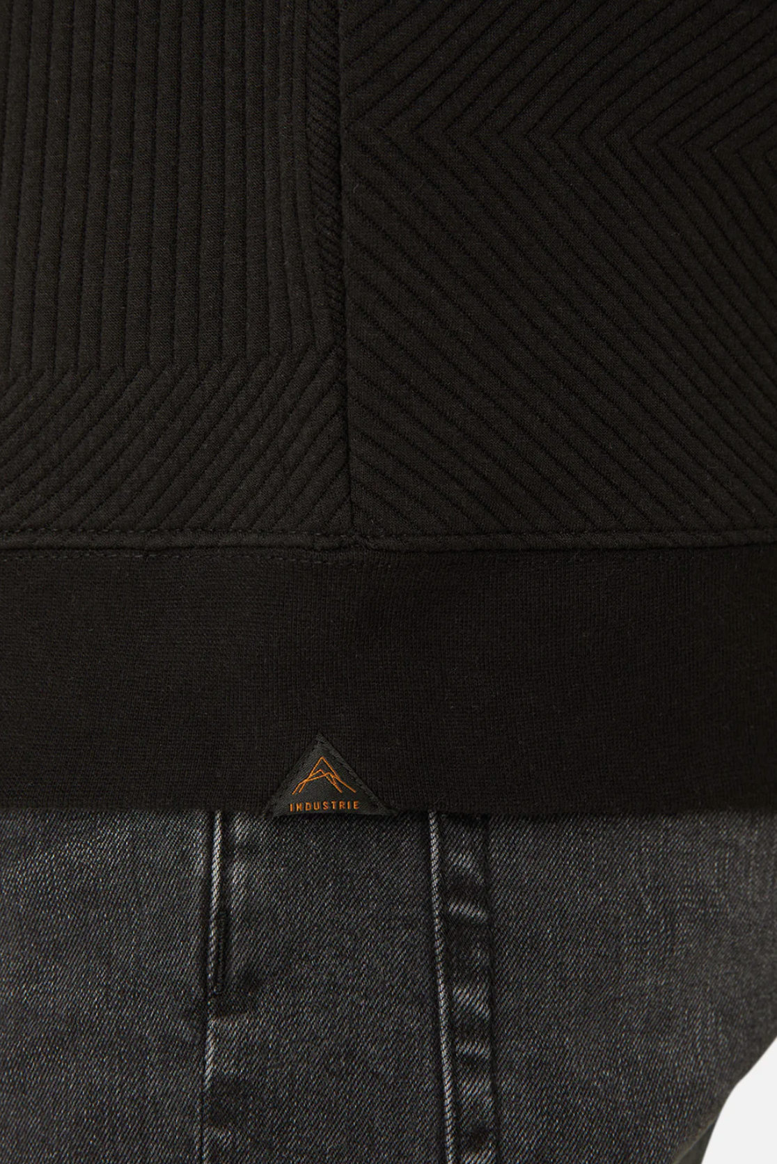 Close-up of a person wearing the Industrie The Robinson Hoodie, a black textured sweatshirt with a relaxed fit. It features a small triangular logo and "INDUSTRIE" near the bottom edge, paired with dark gray denim jeans.