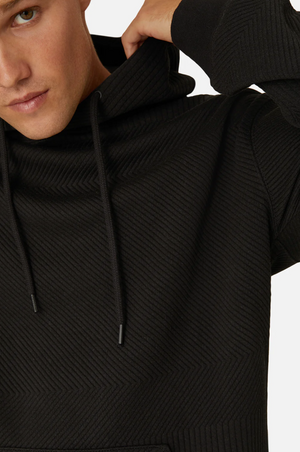 A person wearing the Industrie The Robinson Hoodie, which is black with a textured design and drawstrings. Only a part of their face is visible, showing a serious expression. This hoodie features long sleeves and a relaxed fit, blending comfort with style.
