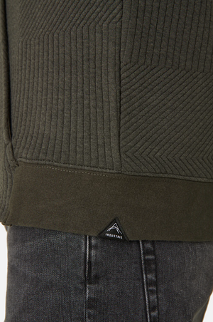 A close-up shows a person in Industrie The Robinson Hoodie, a textured dark green jacket with diagonal patterns and a relaxed fit. A small triangular INDUSTRIE logo is near the hem, and the bottom overlaps dark denim jeans for casual sophistication.