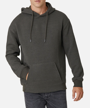 Wearing the Industrie The Robinson Hoodie in dark gray, this person showcases a relaxed fit with a textured design, complete with a front pocket and drawstrings. Paired with dark jeans, the outfit contrasts sharply against the plain white background.