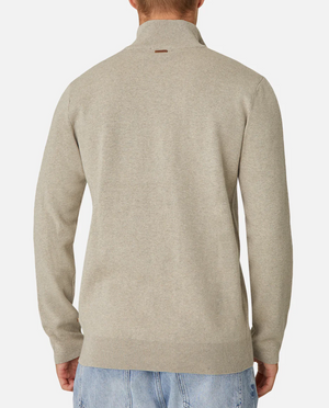 A person wears an Industrie The Lakewood Zip Neck, a beige jumper with long sleeves, ribbed neckline, cuffs, and hem made from 100% cotton. It features a small back tag. They're paired with light blue jeans against a plain white background.