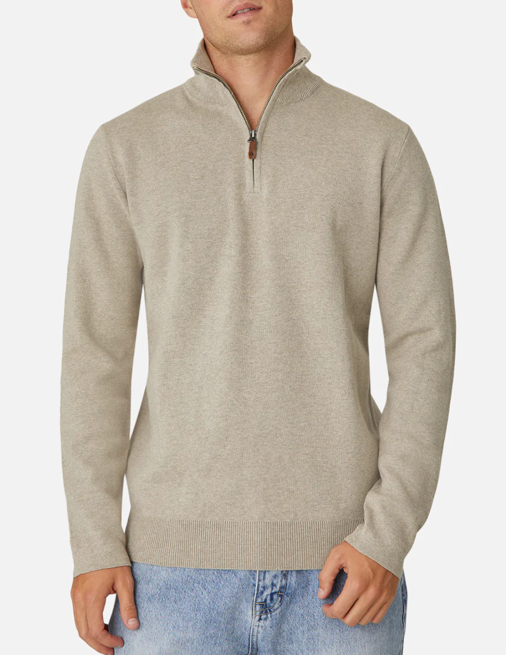 An individual models the Industrie The Lakewood Zip Neck, a beige 100% cotton quarter-zip jumper with a high collar and partially undone zipper, paired with light blue jeans, set against a plain white background. Their left hand gently holds the sweater's hem.