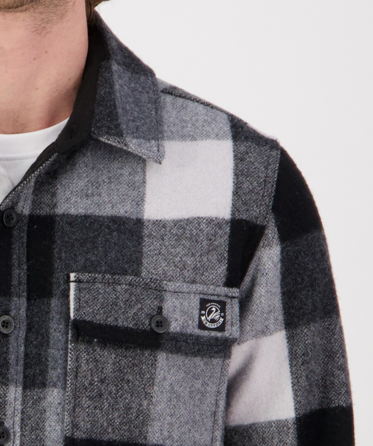 A close-up of someone wearing the Swanndri Kiaraki v3 Wool Shacket in black, gray, and white, featuring a visible front pocket with button and a small logo tag. The person's face is partially out of frame.