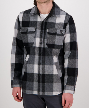 A person wearing a black and white checkered Swanndri Kiaraki v3 Wool Shacket with front pockets and a small chest logo, paired with dark pants, stands against a plain white background.