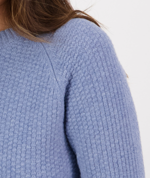 Close-up of an individual wearing a light blue Swanndri Hayes Park Waffle Knit Crew. Only part of the neck, shoulder, and upper arm are visible. The sweater has a ribbed collar and a textured pattern. The background is white.