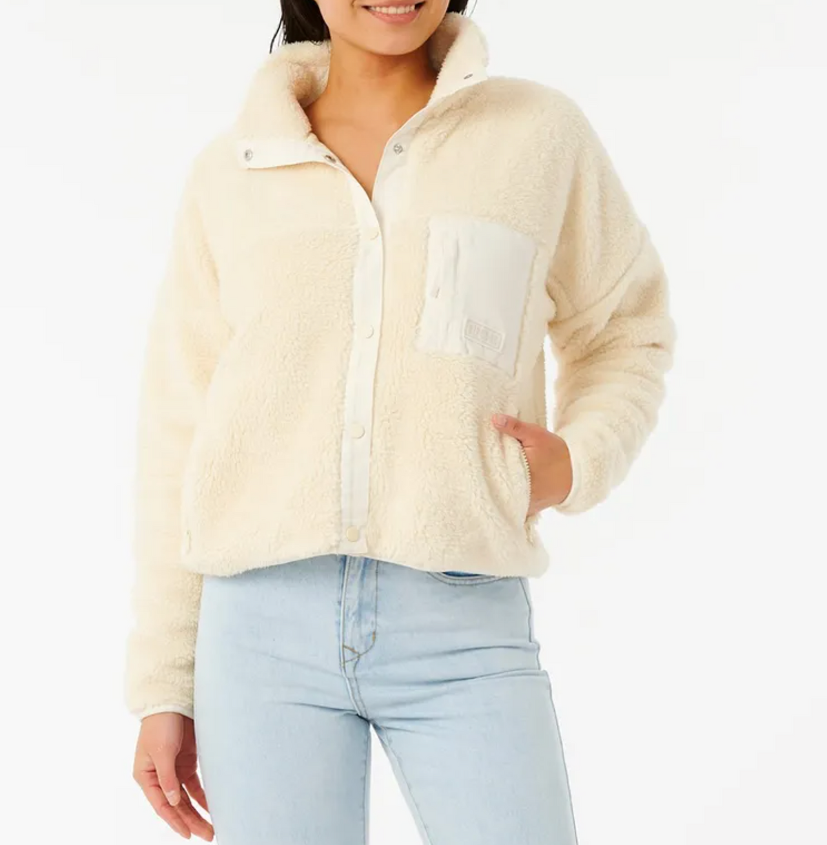 A person in a cream-colored Rip Curl Supply Button Up Polar Fleece with a large front pocket and light blue jeans exudes cozy vibes. Their left hand rests in the pocket against a plain white background.