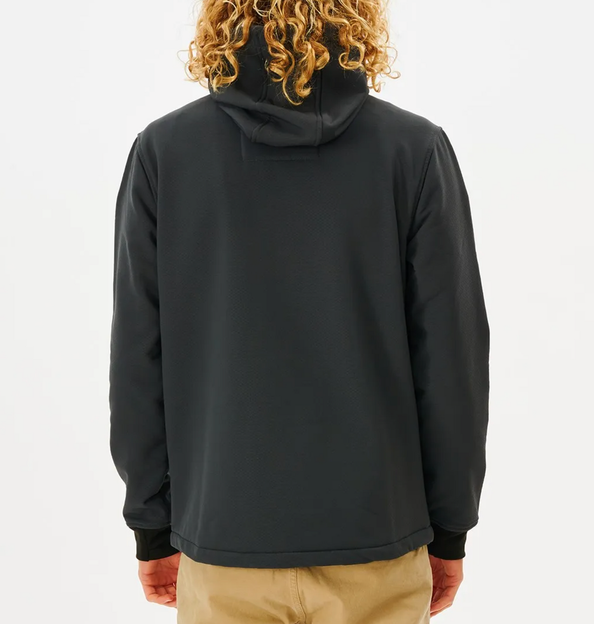 A person with curly blonde hair wears a black Rip Curl Anti Series Soft Tech Fleece Jacket, celebrated for its wind resistance, paired with beige pants, standing back to the camera against a plain white background.