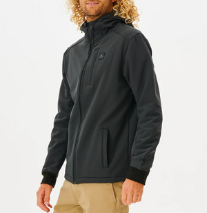 A person with curly hair, wearing a Rip Curl Anti Series Soft Tech Fleece Jacket known for wind resistance, stands against a white background in khaki pants. The dark jacket has a high collar, zippered pockets, and a small chest logo. They look to the side.