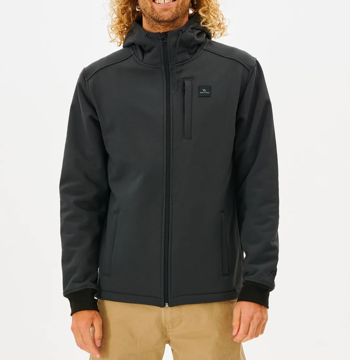 A person with curly hair smiles against a white background, wearing beige pants and the black Rip Curl Anti Series Soft Tech Fleece Jacket featuring wind resistance, a logo, zip-up style, two side pockets, and one chest pocket.