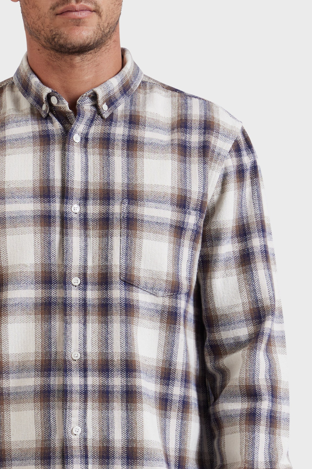 A close-up image of a person wearing the stylish Academy Brand Colorado Shirt, a long-sleeve plaid button-up in shades of white, blue, and brown. Made from 100% cotton, the shirt features a front pocket on the left side. The person is facing forward with their face only partially visible against a plain white background.