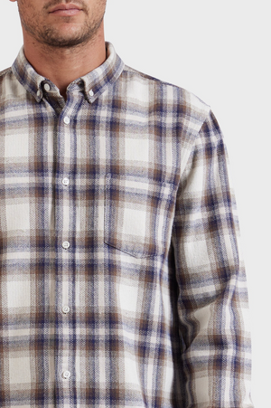 A close-up image of a person wearing the stylish Academy Brand Colorado Shirt, a long-sleeve plaid button-up in shades of white, blue, and brown. Made from 100% cotton, the shirt features a front pocket on the left side. The person is facing forward with their face only partially visible against a plain white background.