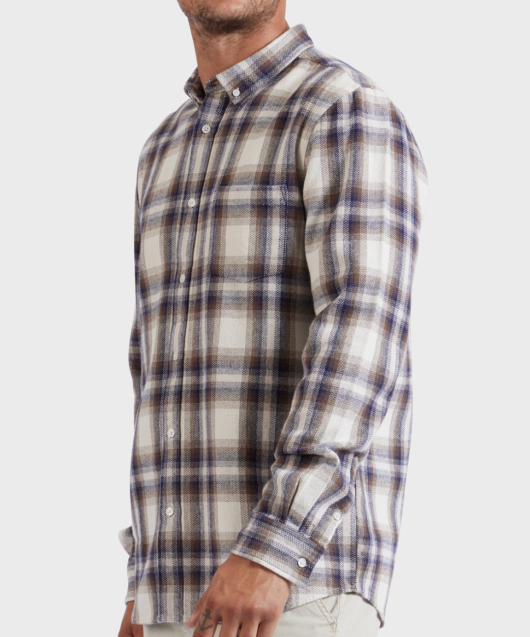 A man is wearing the Academy Brand Colorado Shirt, a stylish long-sleeved button-up with a plaid pattern in shades of blue, beige, and white. Made from 100% cotton, the shirt features a collar and buttoned cuffs. He stands with his left hand partially visible, while his head is cropped out of the image.