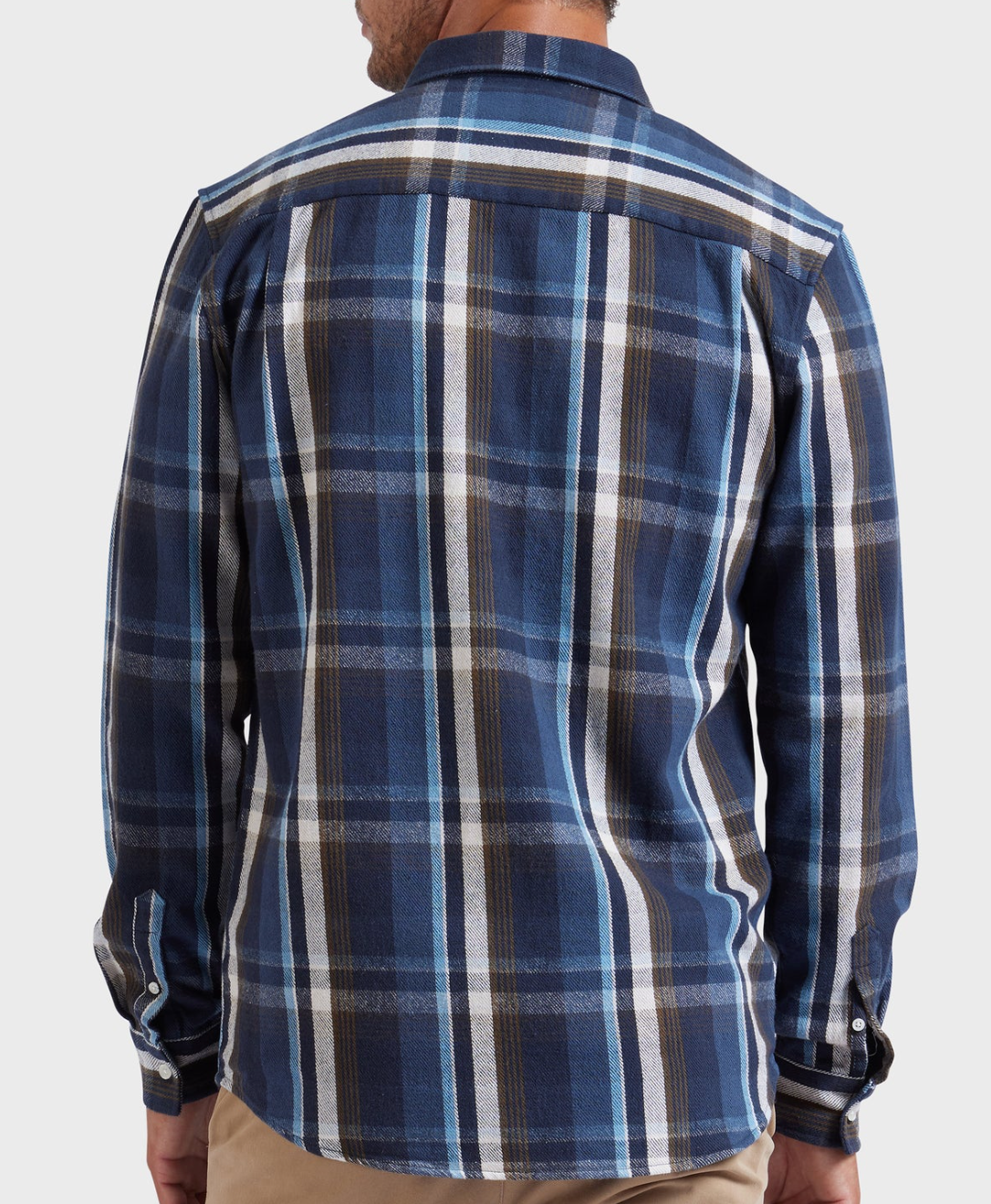A person is shown from the back wearing an Academy Brand So Cal Shirt, a long-sleeved blue plaid shirt with white, black, and brown stripes made from 100% cotton, paired with beige pants against a plain light background.