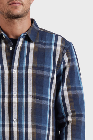 A man wears the Academy Brand So Cal Shirt, a blue, white, and brown plaid 100% cotton button-up, shown from shoulders to mid-chest against a plain light background with only the lower half of his face visible.