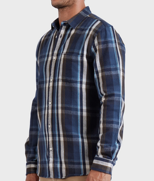 A man stands against a plain light background wearing the Academy Brand So Cal Shirt, a long-sleeve, button-up plaid shirt made from 100% cotton in blue, white, and brown with a collar and chest pocket.
