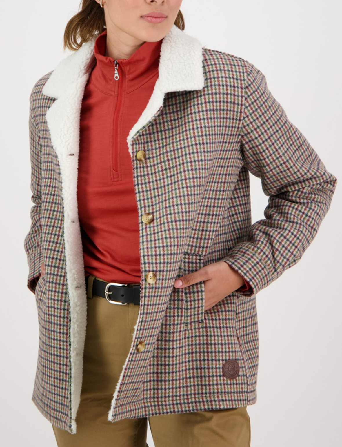 A person in a red zip-up top, tan pants, and a Swanndri Capitola Sherpa Jacket with brown buttons, side pockets, plaid wool/merino fabric, and a white sherpa collar stands with their hands in their pockets.