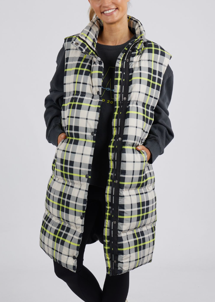 A person stands against a plain background wearing the Foxwood Check It Long Line Vest, featuring white, black, and lime green checks. They are dressed in a dark long-sleeve shirt and dark pants underneath the vest.