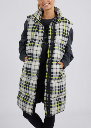 A person stands against a plain background wearing the Foxwood Check It Long Line Vest, featuring white, black, and lime green checks. They are dressed in a dark long-sleeve shirt and dark pants underneath the vest.